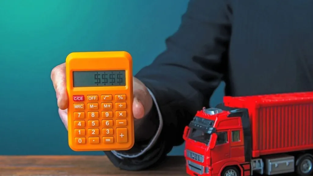 Pessoa segurando calculadora com uma miniatura de caminhão na mesa.